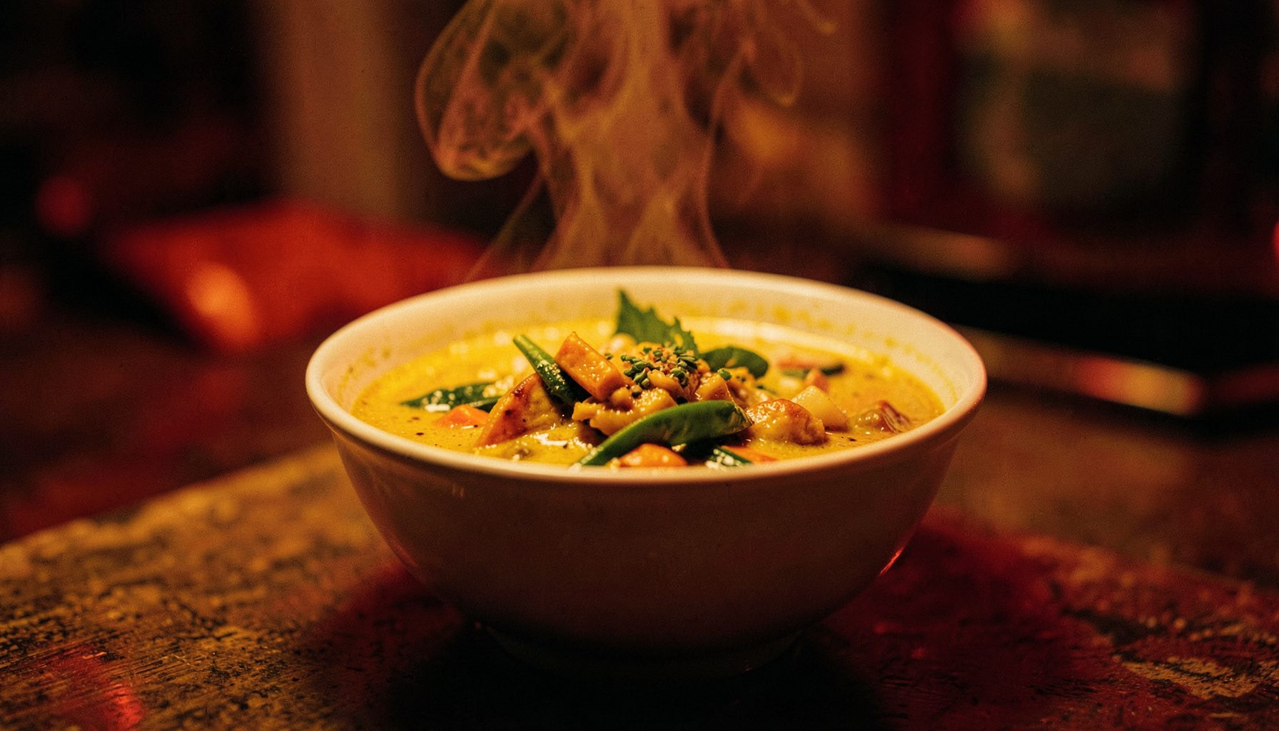 Wide cinematic shot of a steaming bowl of vegan Thai curry with vegetables, shallow focus, deep red and warm mustard lighting, cozy mood
