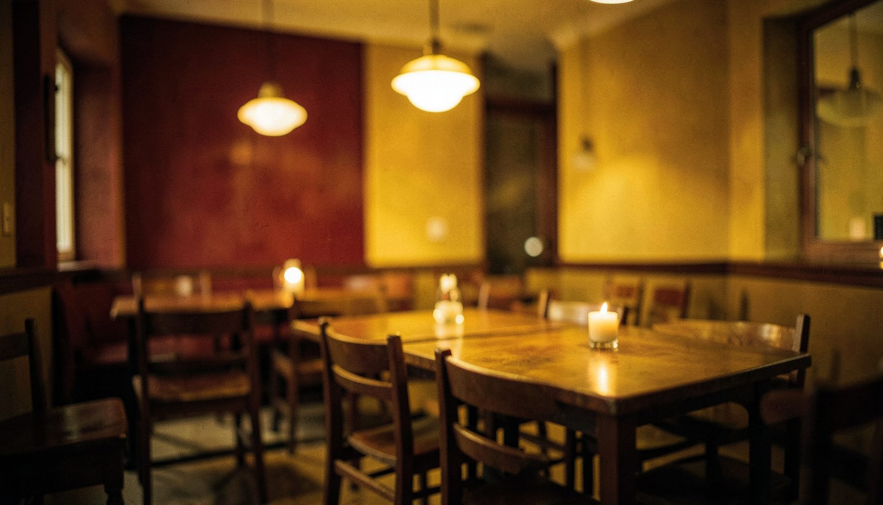 A warm, softly lit dining room with deep red and mustard yellow accents, wooden tables set simply, and a gentle blurred glow from hanging lights creating a cozy, welcoming atmosphere